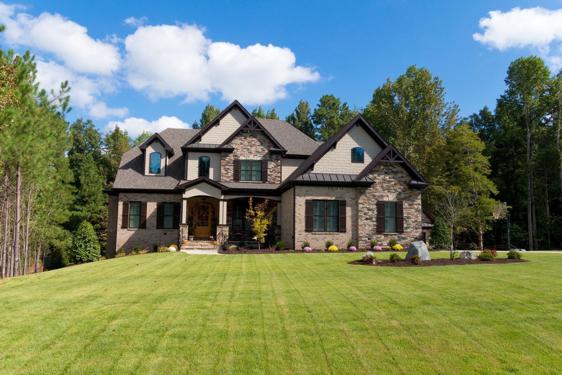 Two-story brick and stone house on a grassy hill, with trees and a blue sky.