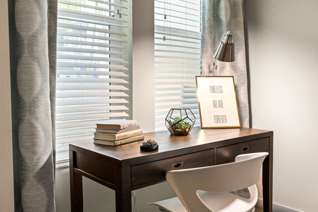 Wooden desk and white chair by a window with horizontal blinds; framed print and terrarium on desk.