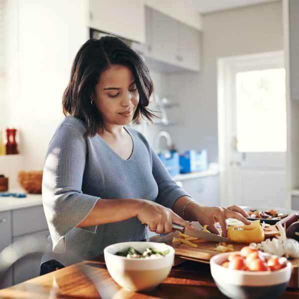 A person in a light blue top preparing fresh vegetables on a wooden cutting board in a sunlit kitchen.