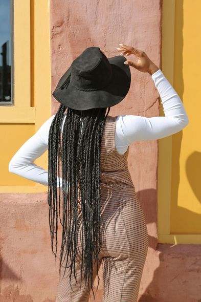 A woman wearing a black hat and overalls is standing in front of a yellow wall.