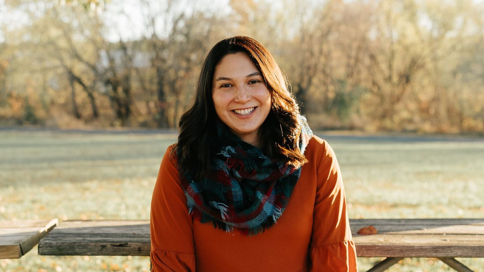 A woman in an orange sweater and scarf is sitting on a picnic table.