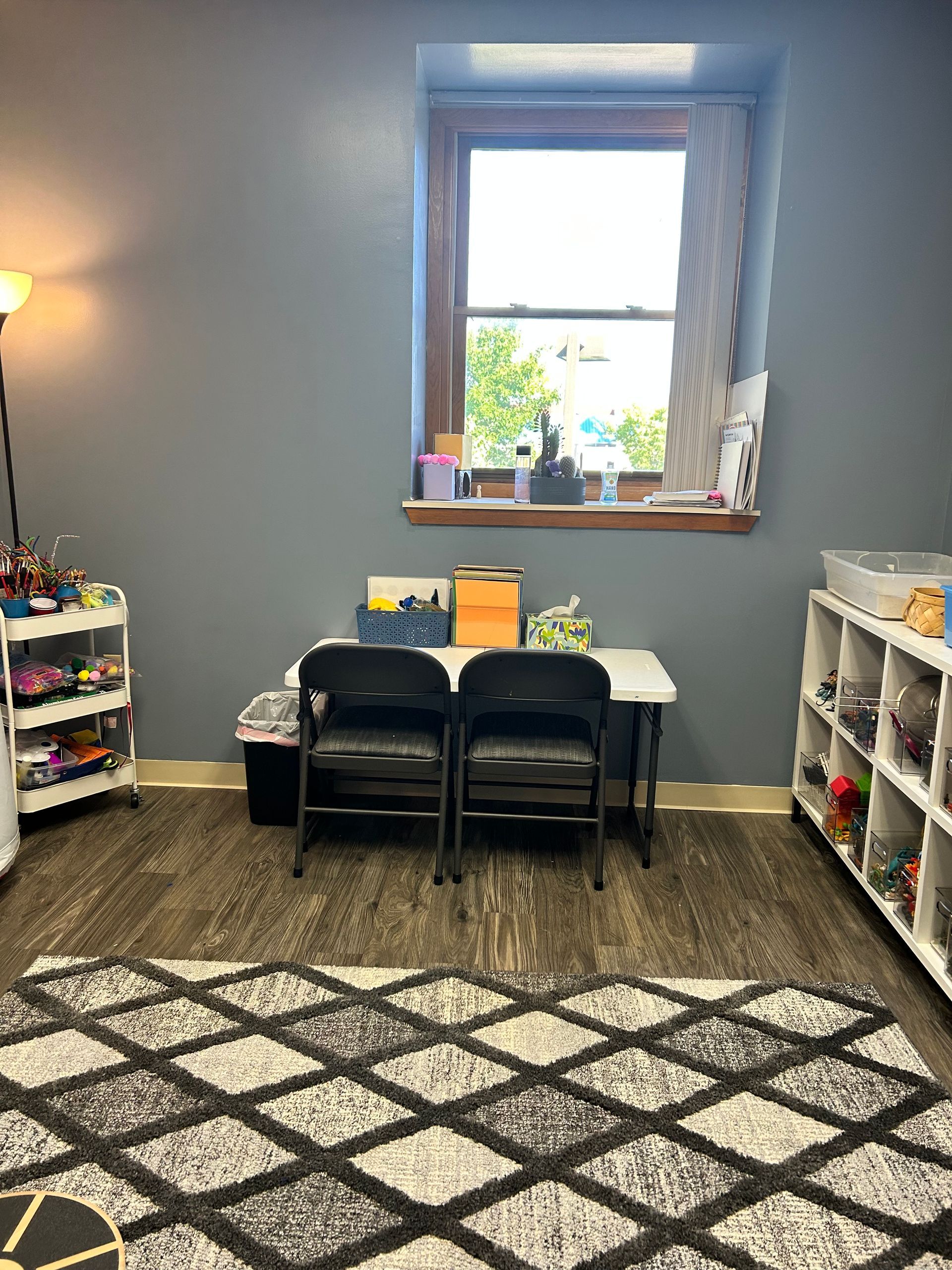 a play area at Peck Counseling with a rug , chairs , a table and a window .