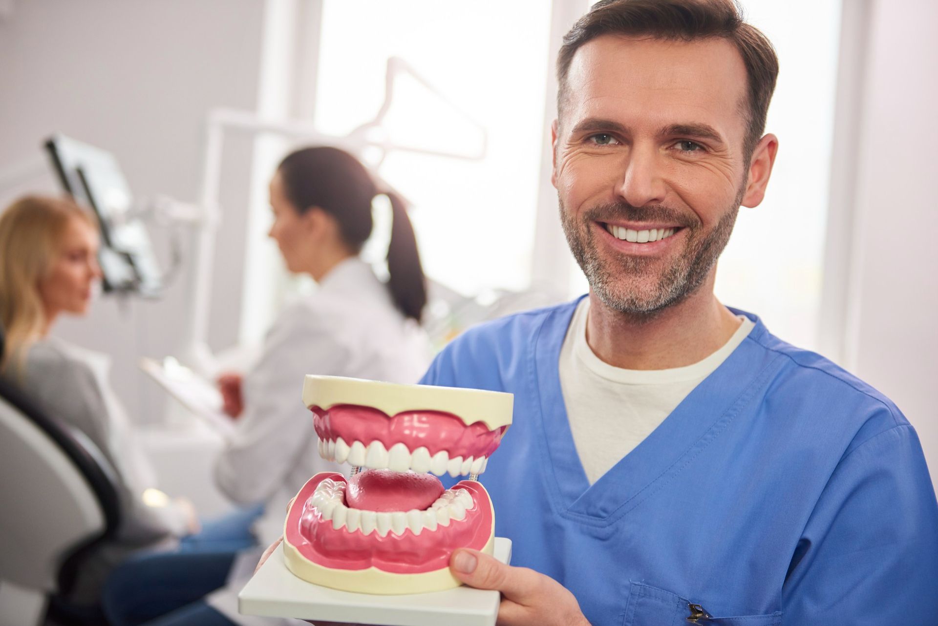 Dentista con un modelo dental, sonriendo en una consulta. Al fondo, un paciente y su asistente.