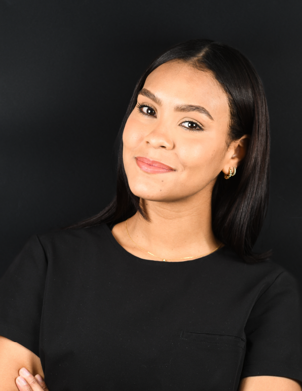 Una mujer de cabello oscuro sonríe, con los brazos cruzados, vistiendo un top negro sobre un fondo negro.
