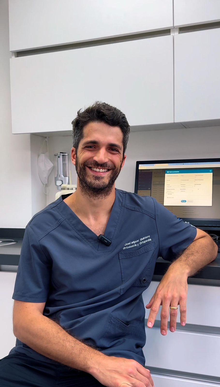 Un hombre con uniforme azul sonríe en una consulta dental. Se sienta junto a una computadora y un mostrador.