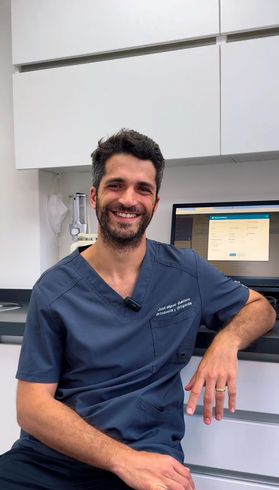 Un hombre con uniforme azul sonríe en una consulta dental. Se sienta junto a una computadora y un mostrador.