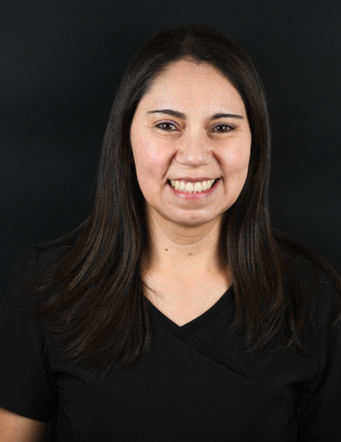 Mujer con cabello largo y oscuro, sonriendo a la cámara, vistiendo un top negro sobre un fondo oscuro.