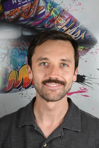 Hombre de cabello oscuro y bigote sonriendo, vistiendo un polo gris, frente a un grafiti.