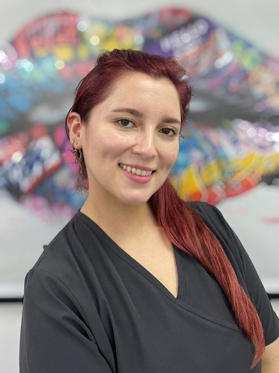 Una mujer de cabello castaño rojizo sonríe, con un top negro. Al fondo, una colorida obra de arte labial.