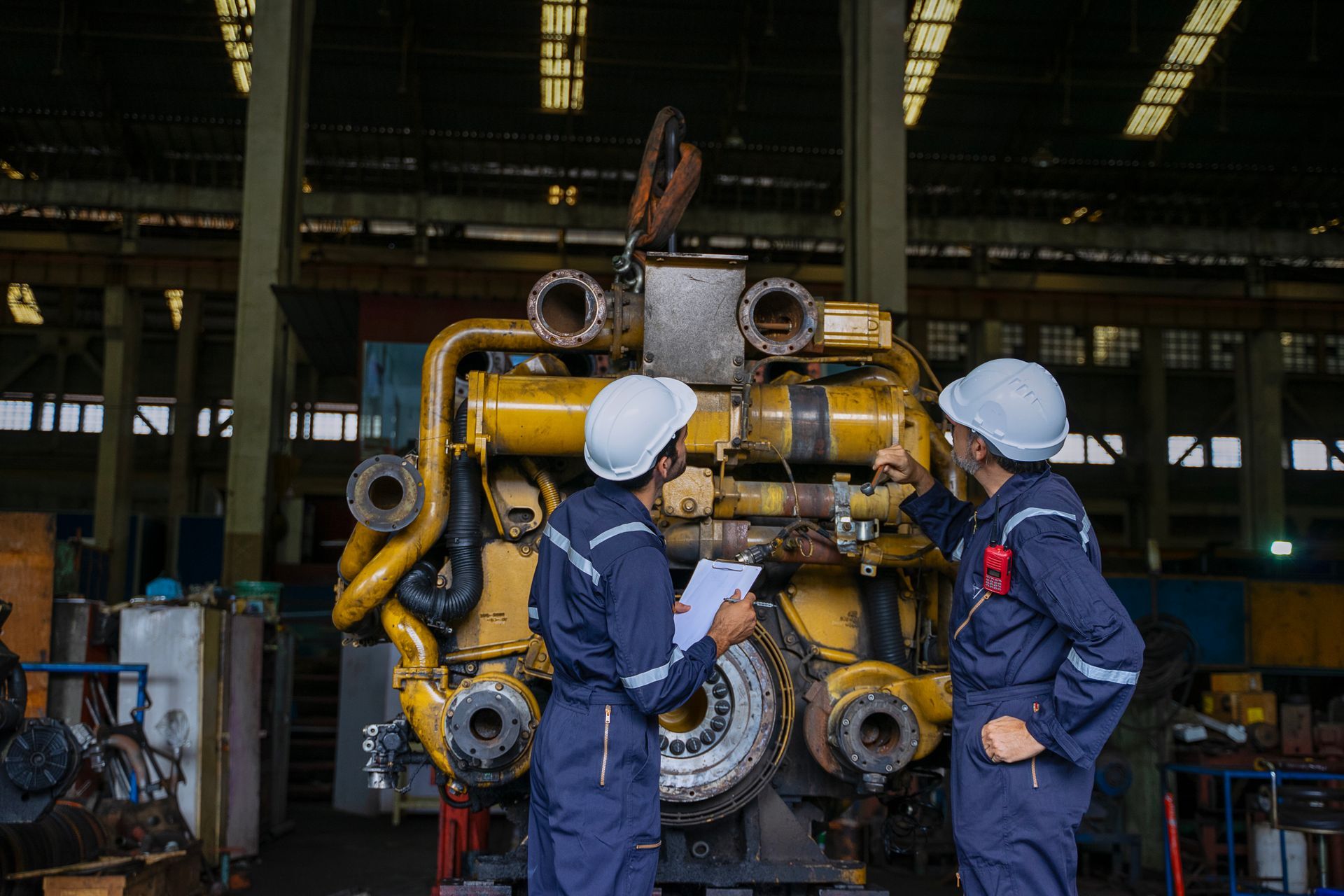Two workers in blue coveralls and white helmets inspect a large engine in a factory.