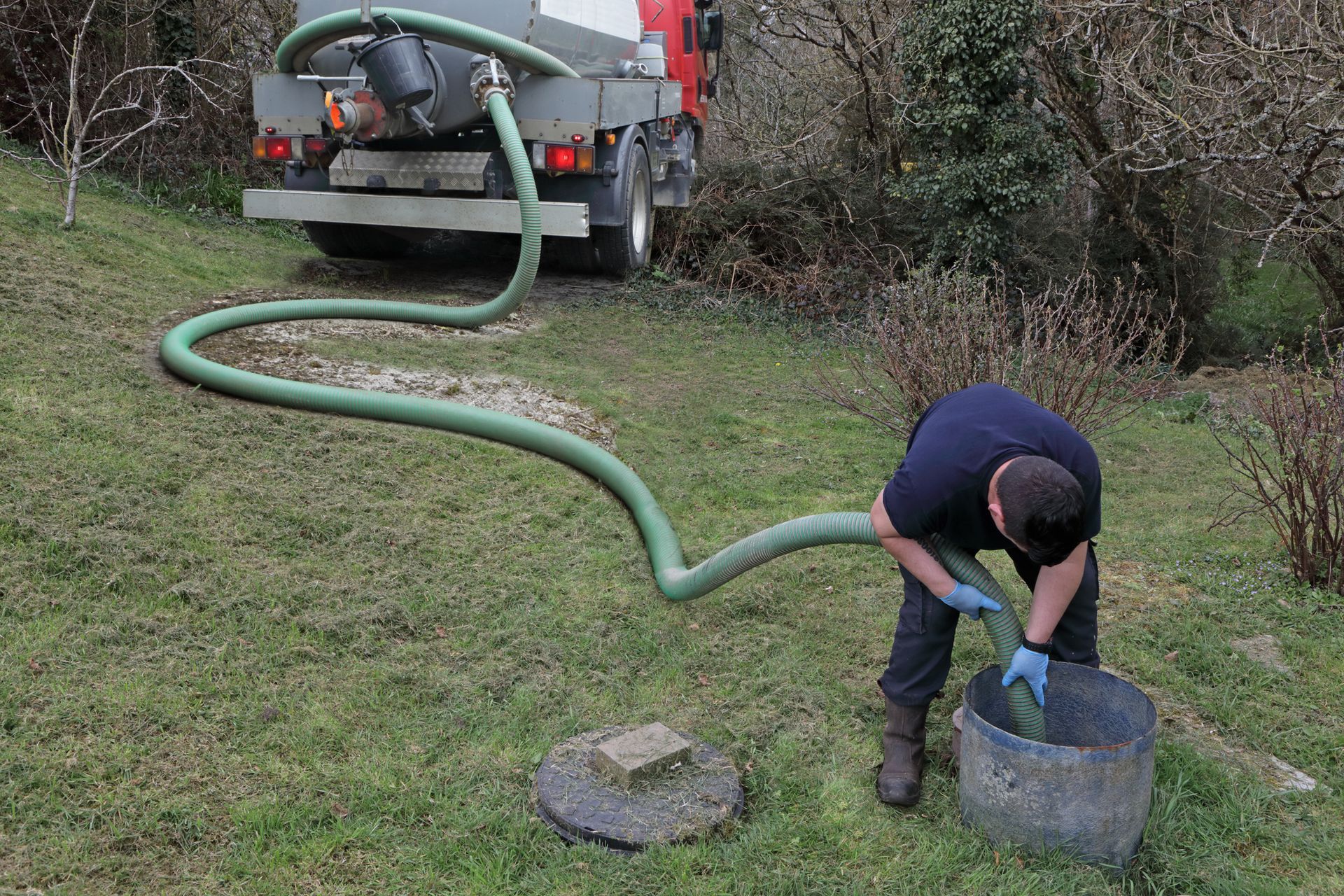 Worker providing septic tank cleaning services with vacuum truck and green hose on grassy field.