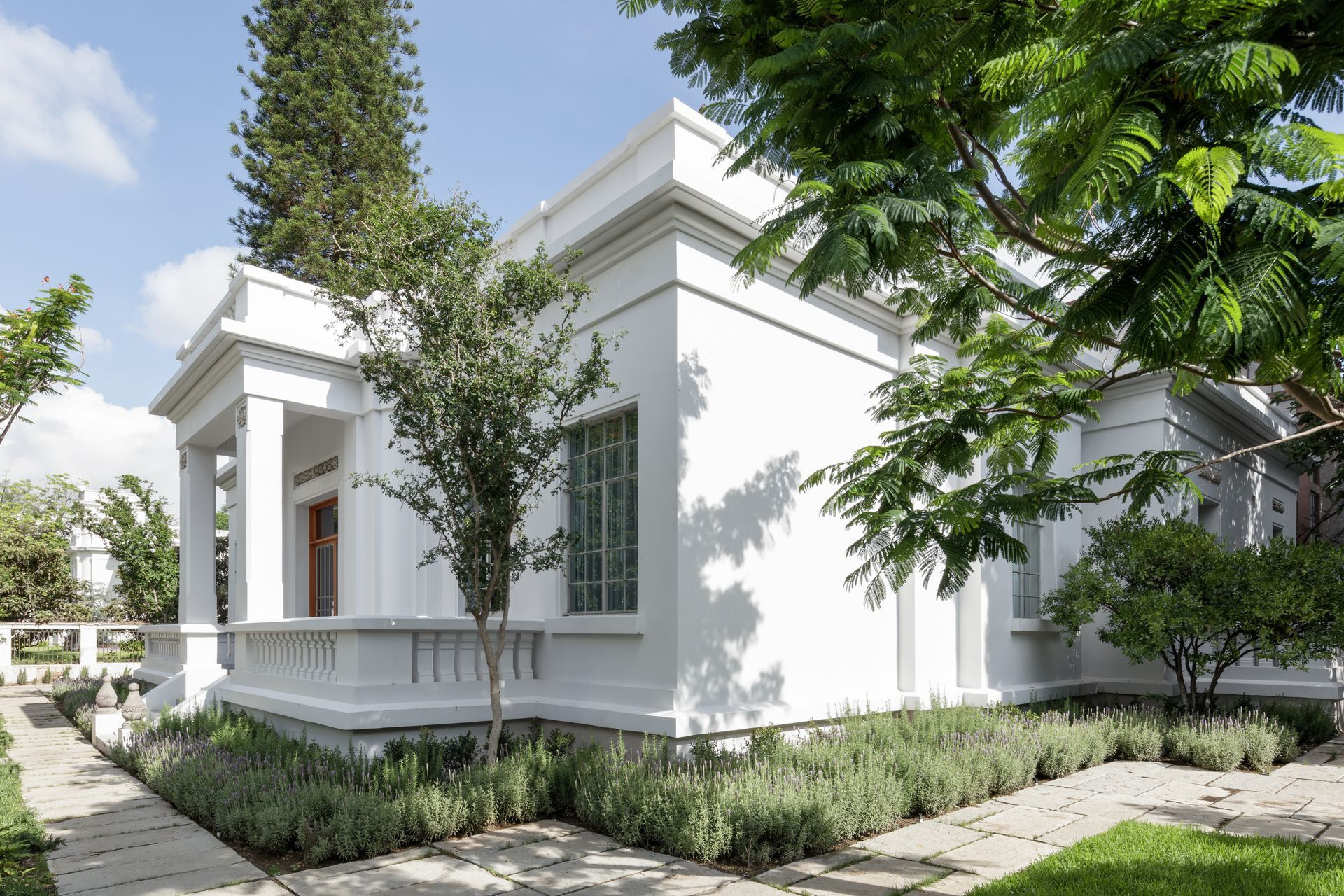 White, colonial-style building with a porch, surrounded by greenery and a stone walkway, on a sunny day.