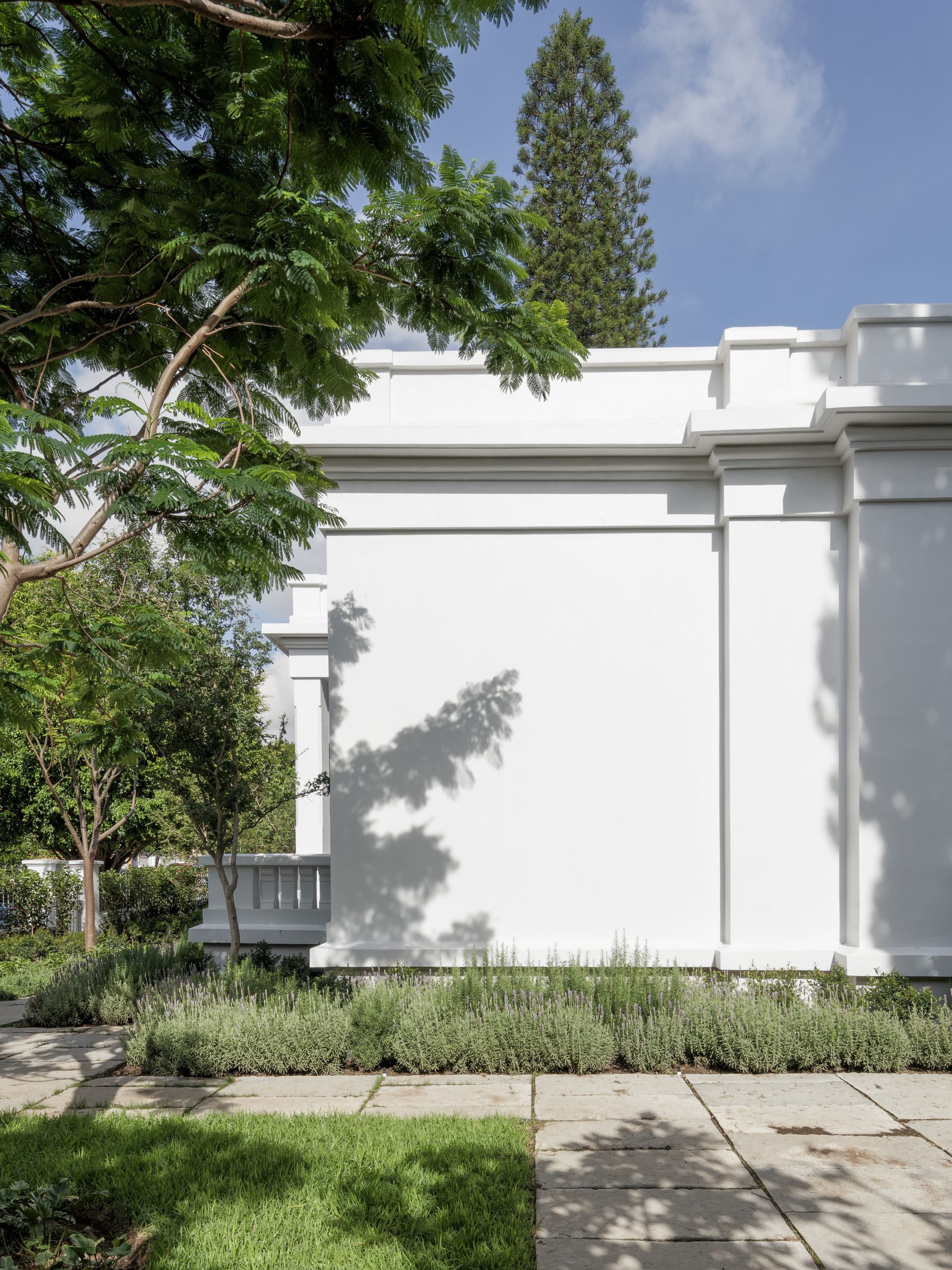 White building facade with tree shadows, surrounded by greenery, against a blue sky.