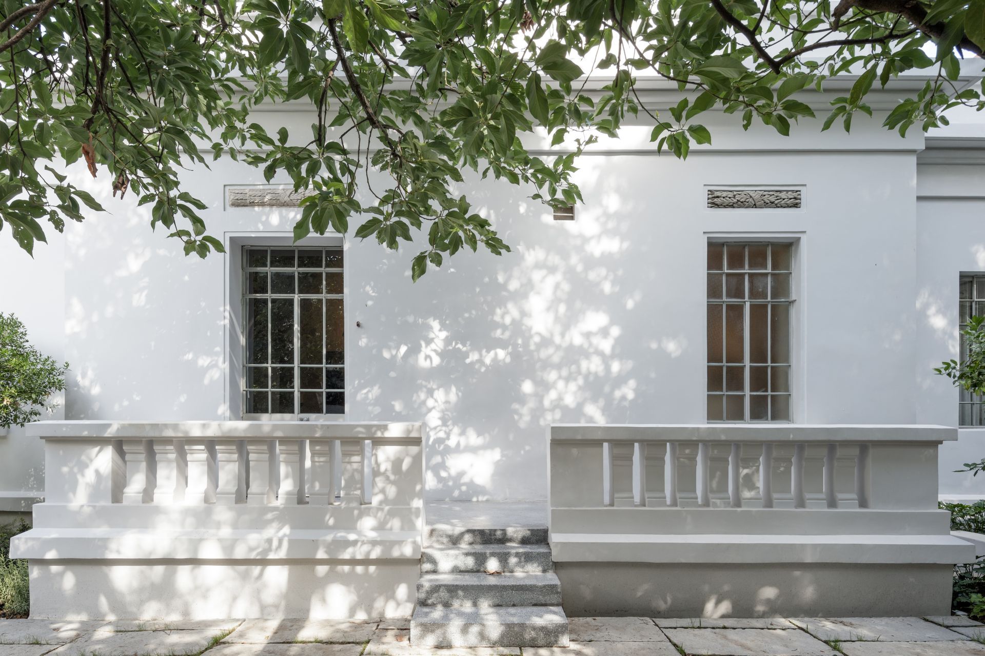 White building with windows and a balcony, shaded by green tree leaves.