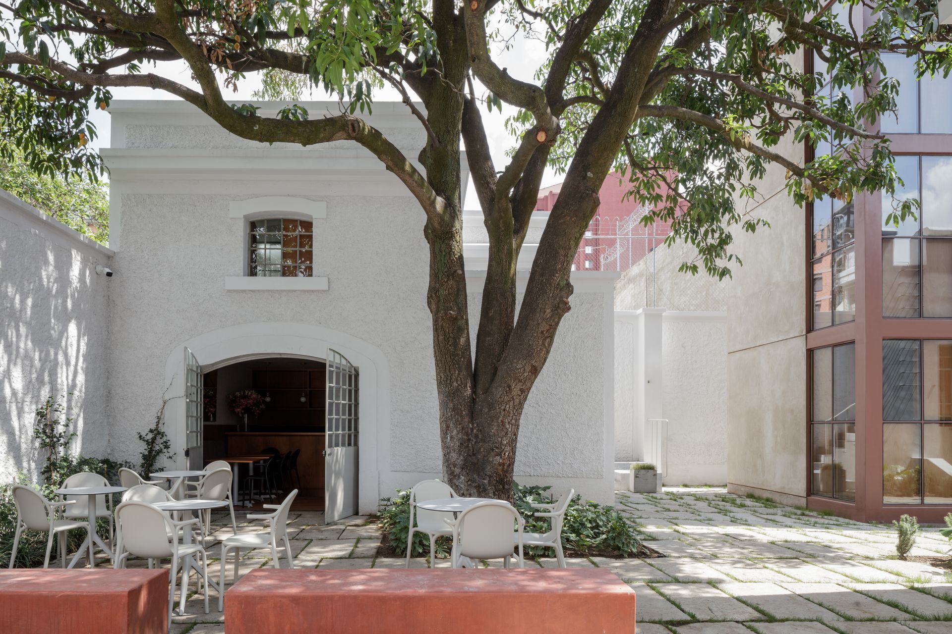 Courtyard with a white stucco building, tree, outdoor seating, and modern glass structure in background.