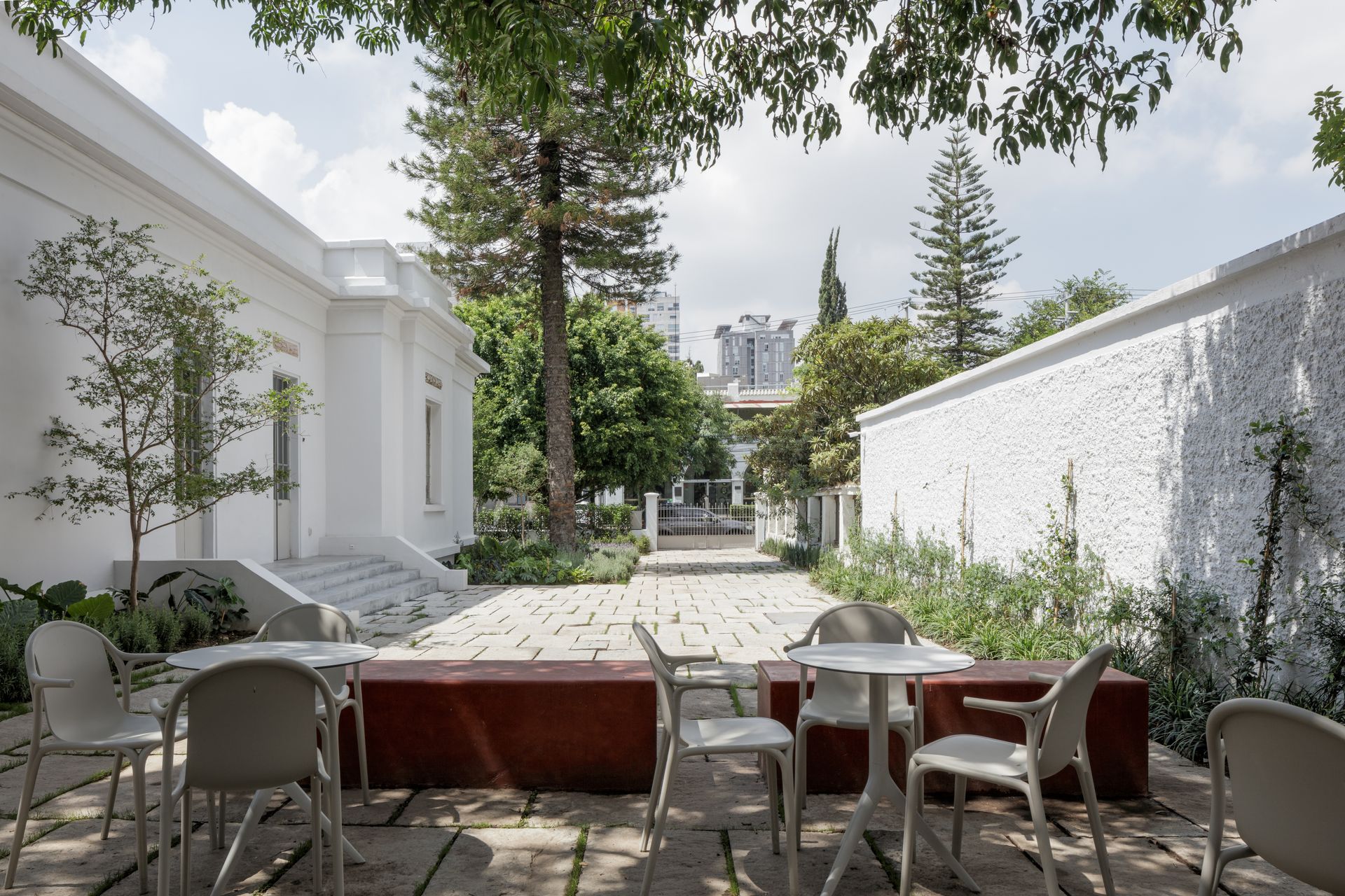Outdoor patio with white walls, red bench, and white chairs. Sunlight filters through trees.