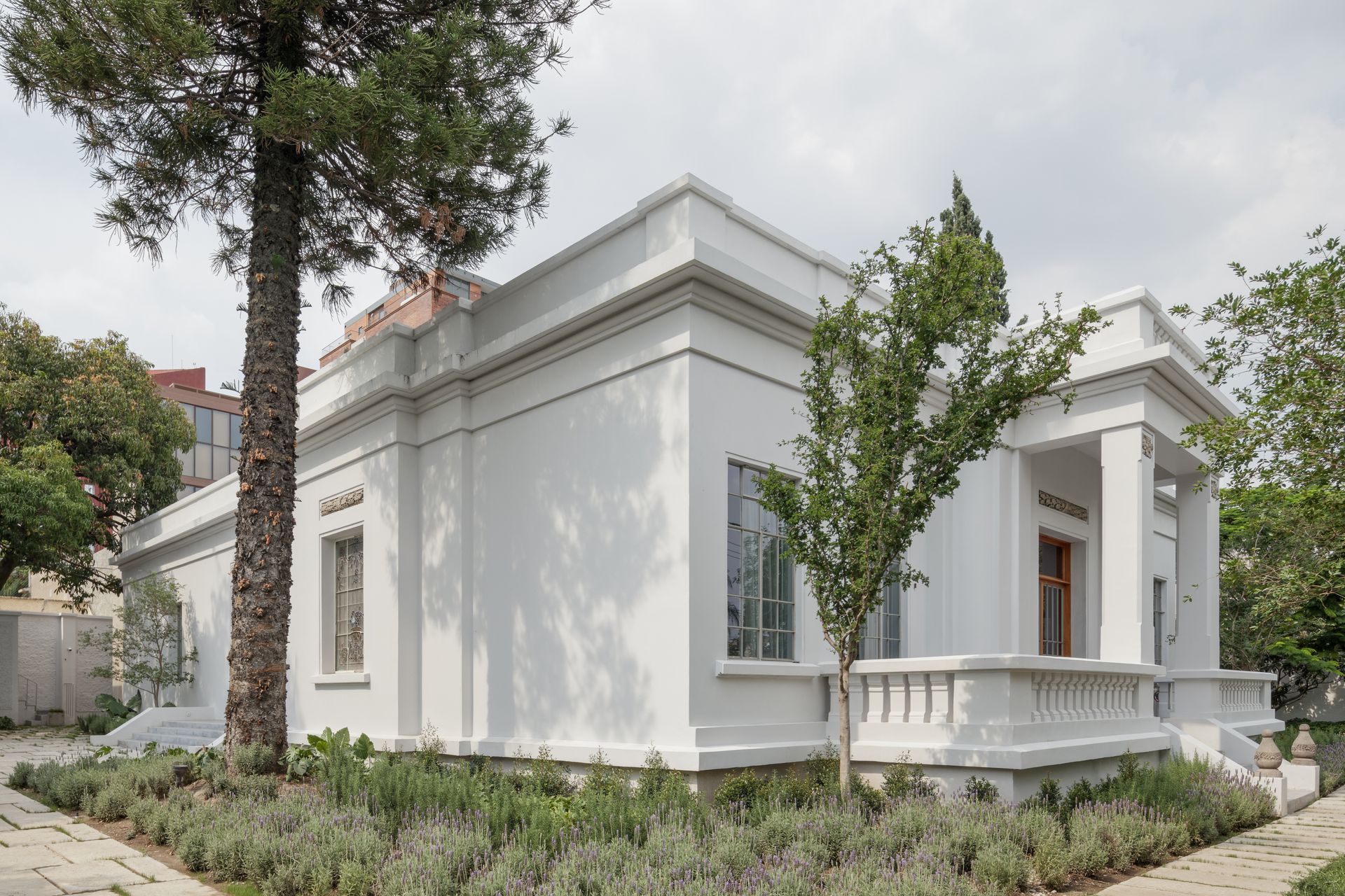 White building with columns, trim, and windows, surrounded by greenery and a tree; daylight.