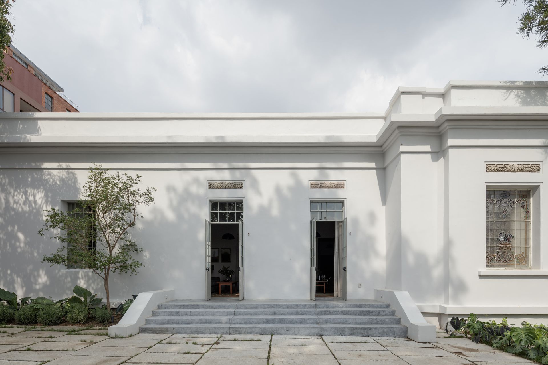 White building with two open doorways, small steps, and foliage.