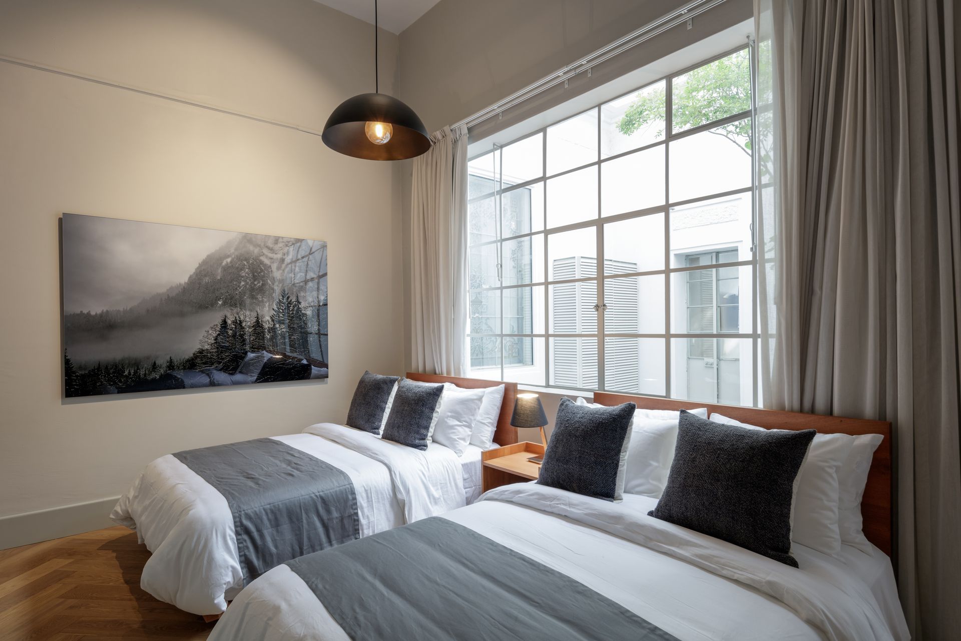 Two beds in a bedroom with gray bedding, large window, artwork, and a hanging light.