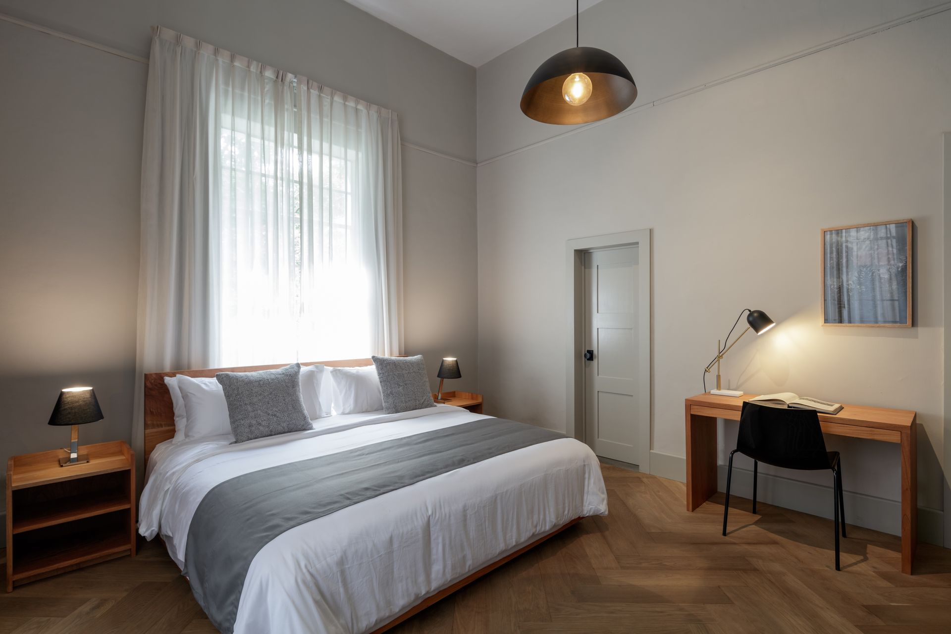 Bedroom with large bed, desk, and lamp near a window. Neutral color scheme with wood accents.