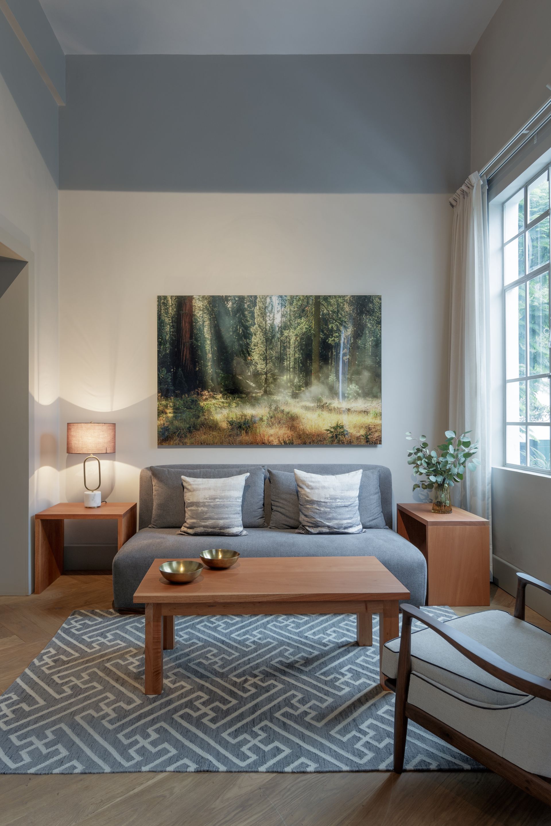 Living room with gray sofa, wooden furniture, artwork of a forest, and patterned rug.