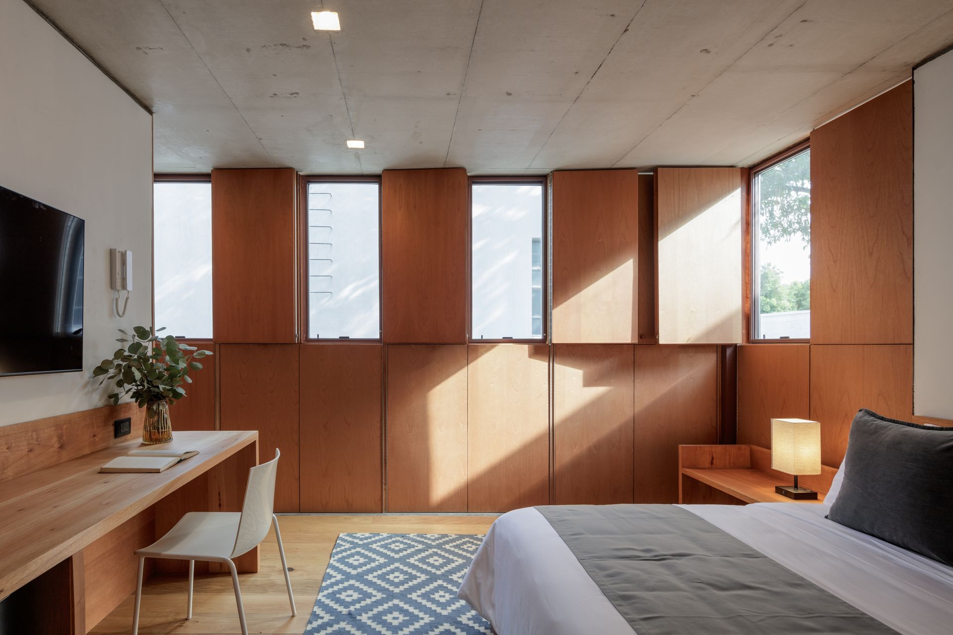 Bedroom with wood paneling, windows, desk, bed with gray blanket, and blue patterned rug.