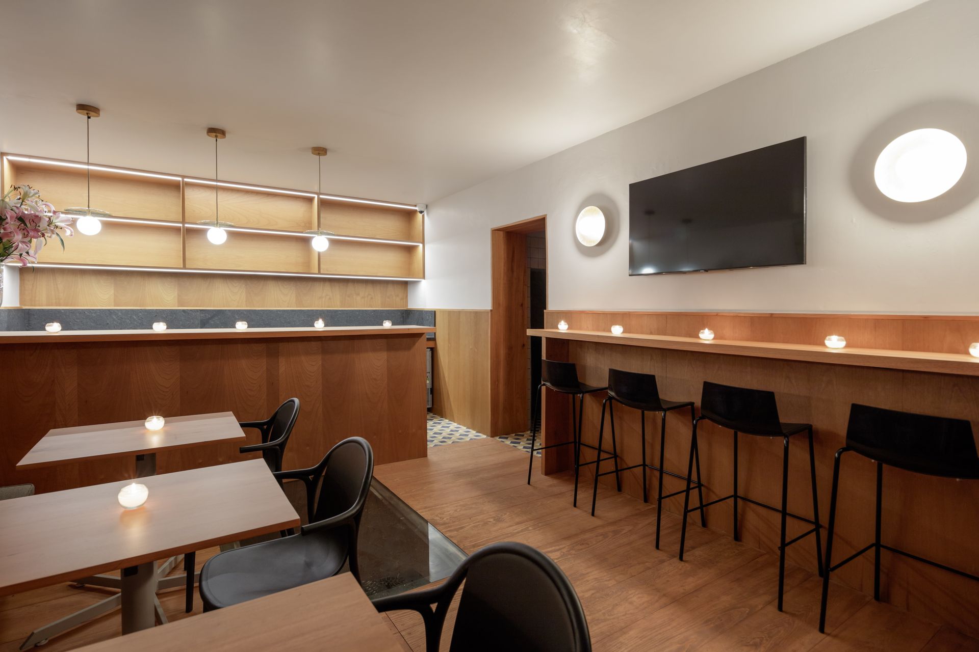 Interior of a bar with wooden counters, shelves, tables, and black stools. There is a television on the wall.