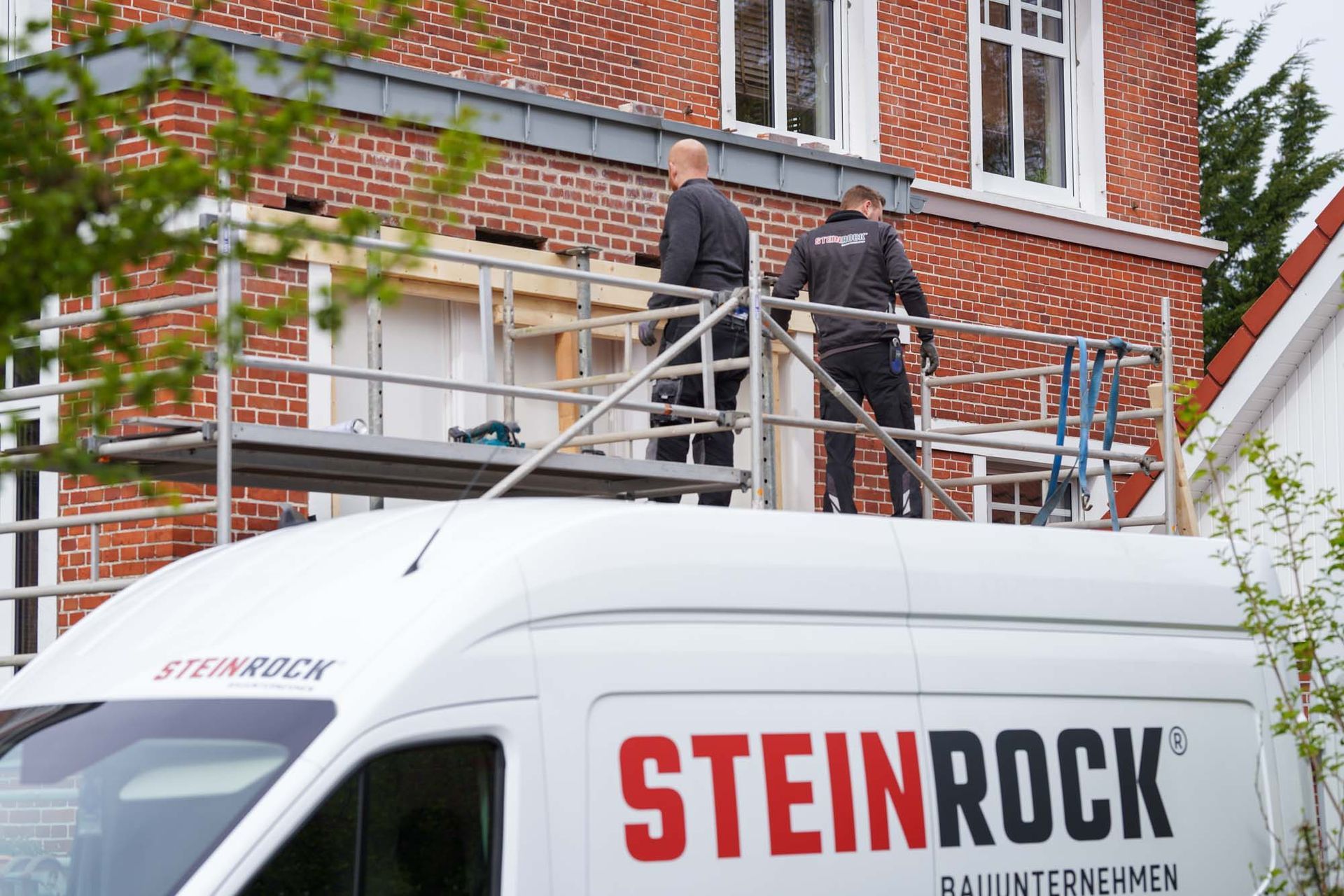 Arbeiter auf einem Gerüst über einem weißen Firmenfahrzeug neben einem Backsteingebäude.