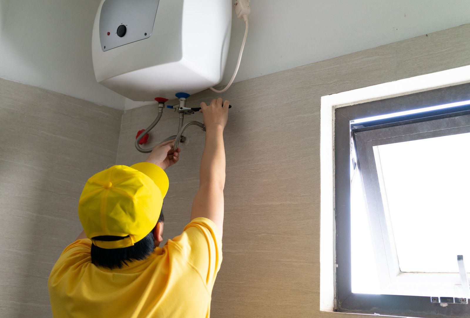 A man in a yellow hat is fixing a water heater in a bathroom.