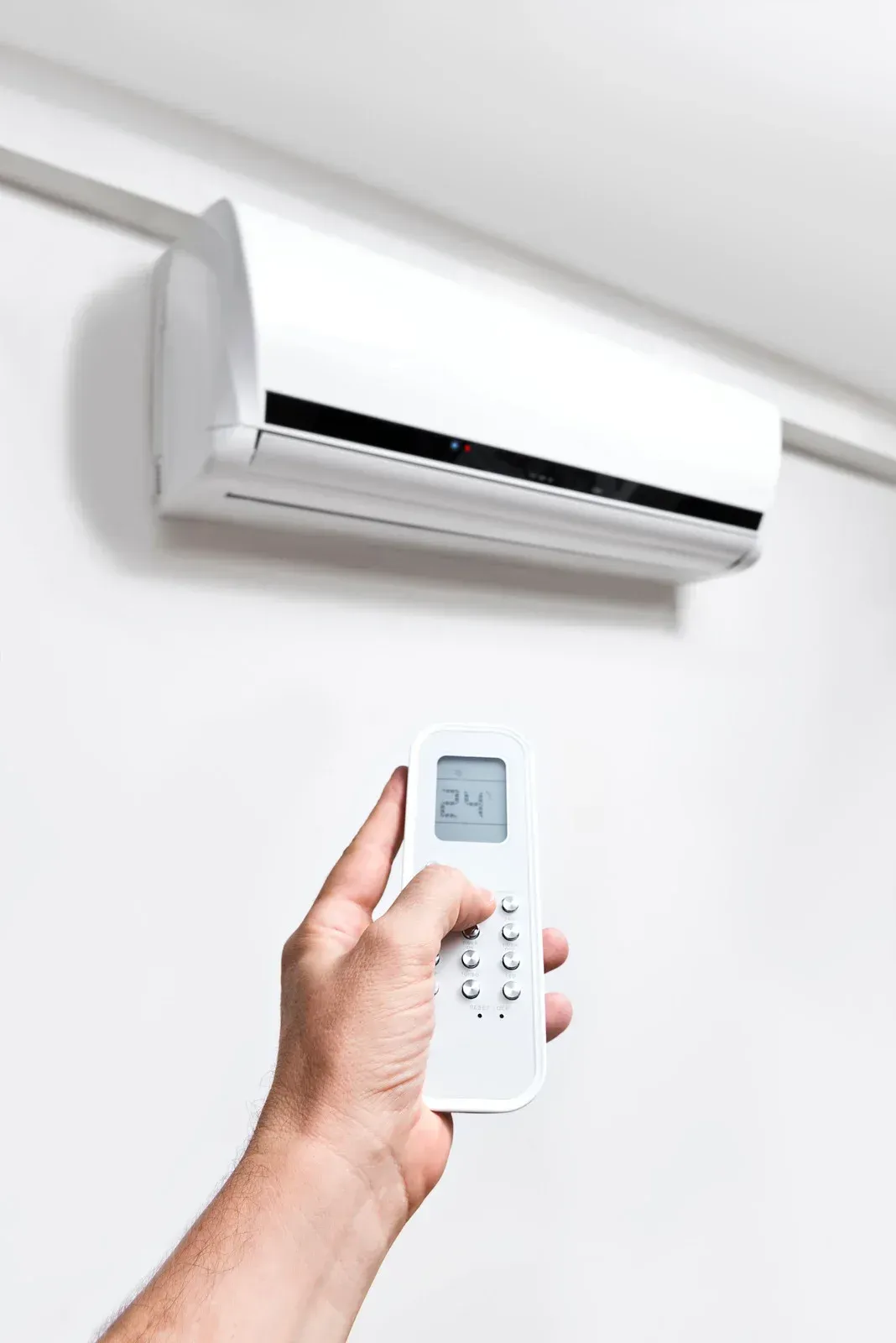 Hand holding a remote control pointing to a white wall-mounted air conditioner. Display shows 24 degrees.