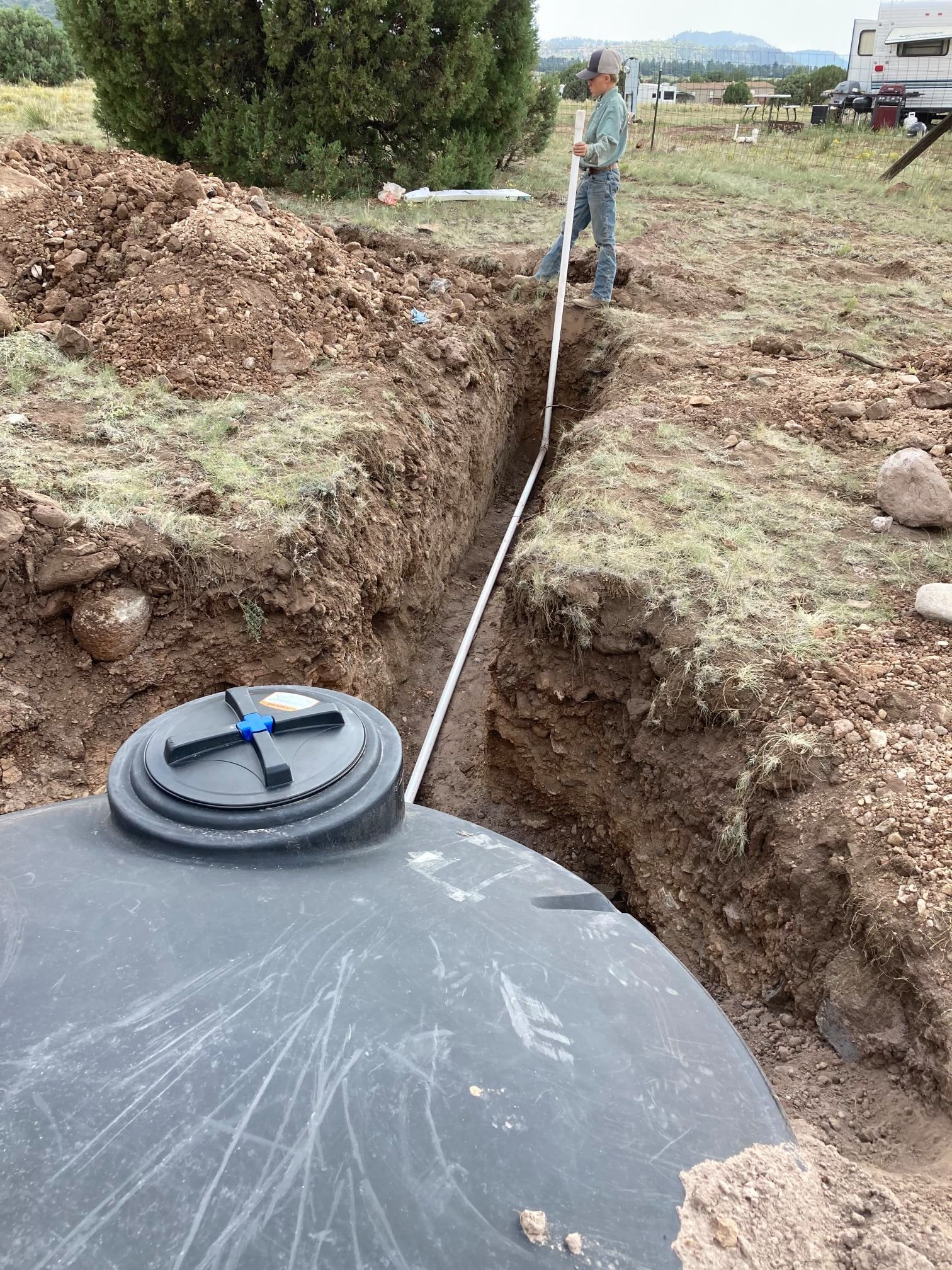 Person laying white pipe in a trench next to a large black water tank outdoors.