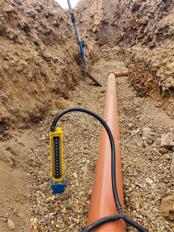 A brown pipe in a trench with a black device and measuring tool, likely for water or utilities.