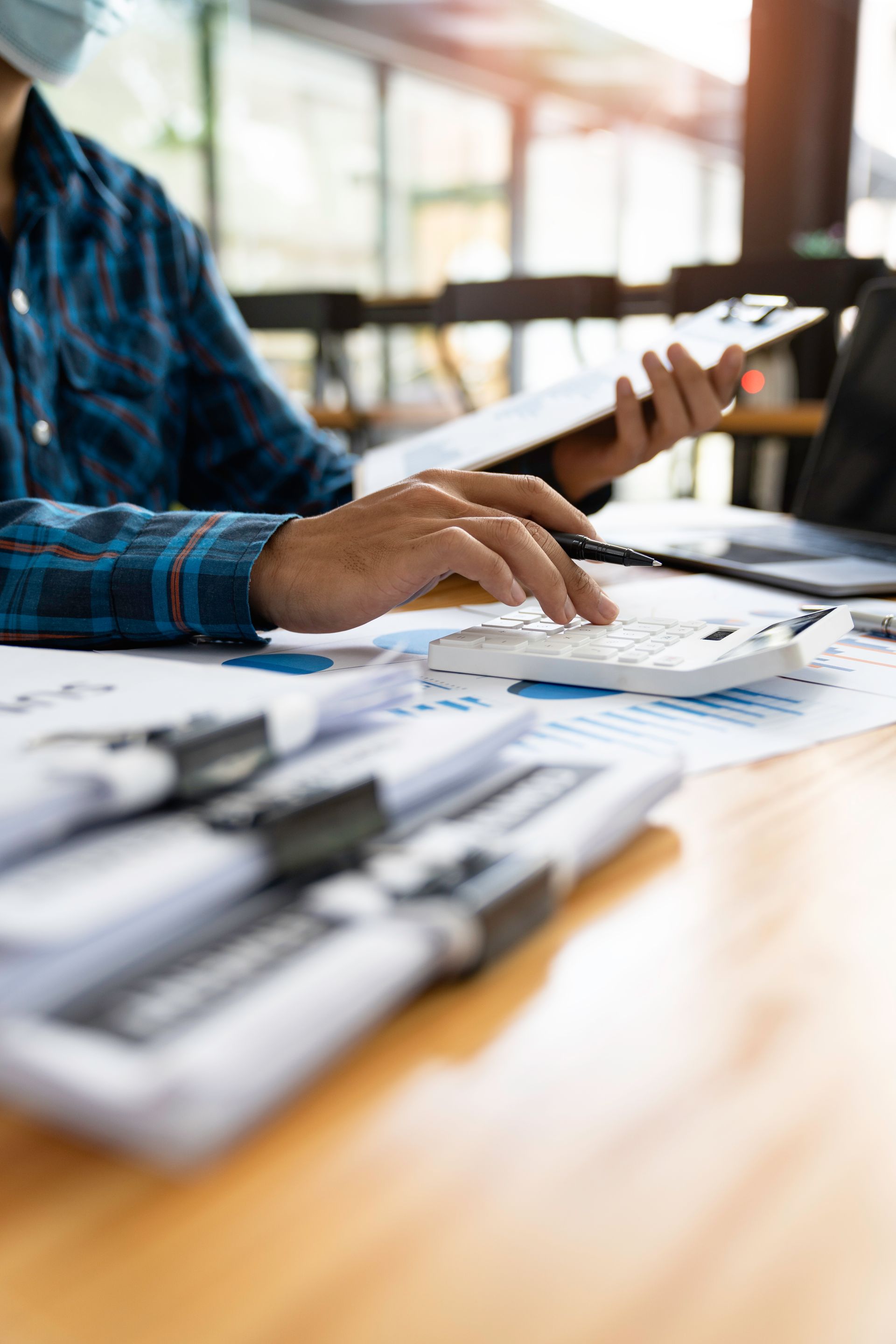 A person in a blue plaid shirt uses a calculator while reviewing papers at a desk with a laptop.