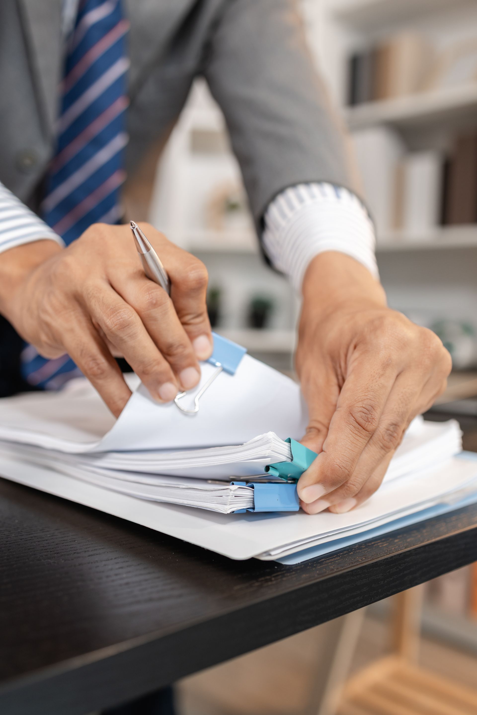 A person in a suit and tie clips documents together with a binder clip while holding a pen at a desk.