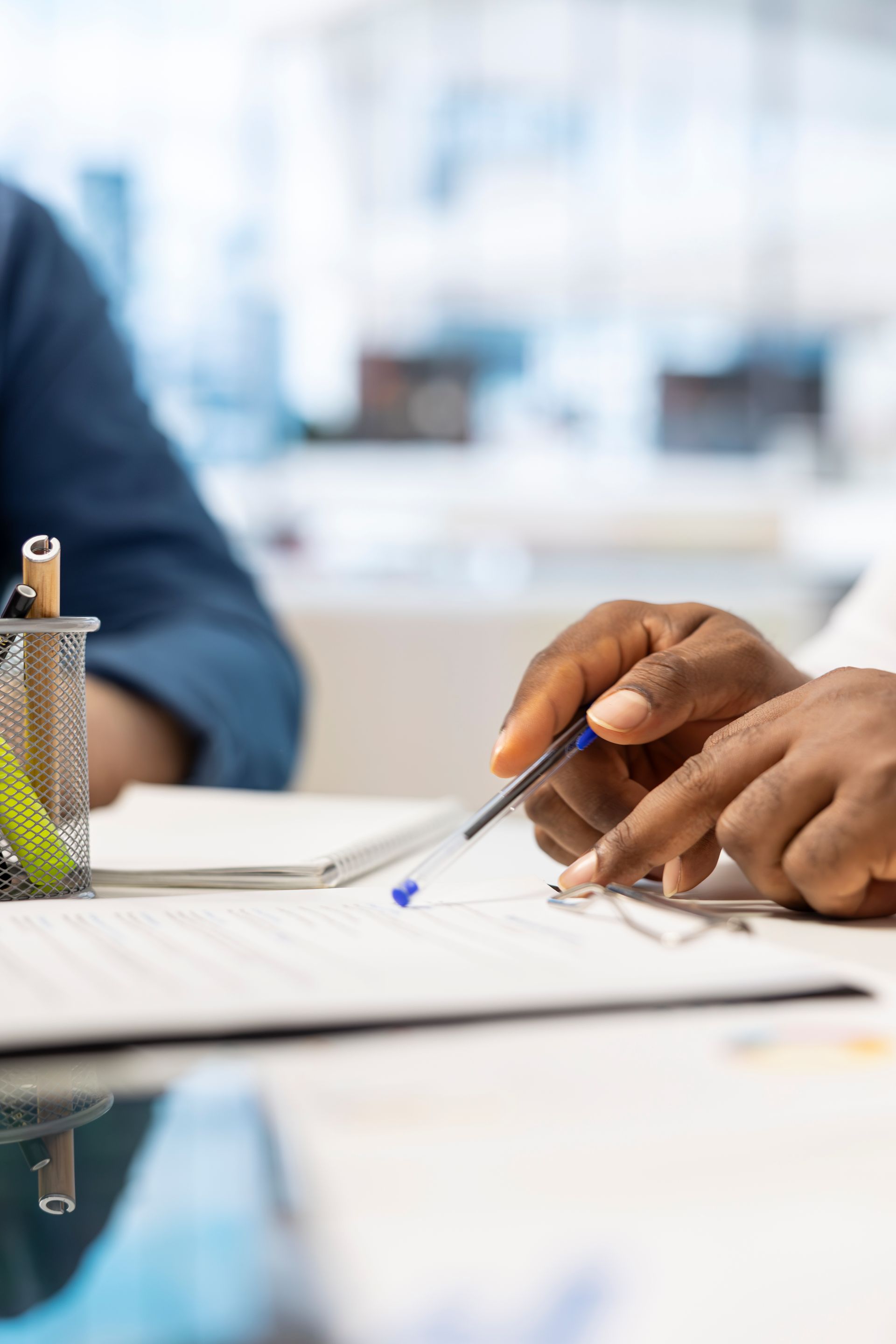 Close-up of hands pointing to a document on a desk with a pen holder nearby, set in a bright office environment.