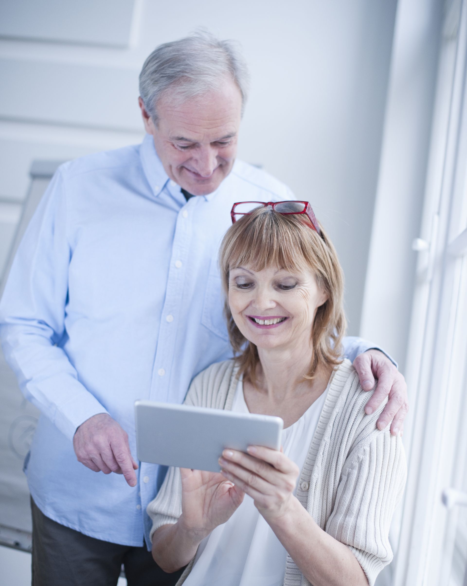 Two people standing near a window looking at a tablet together and smiling.