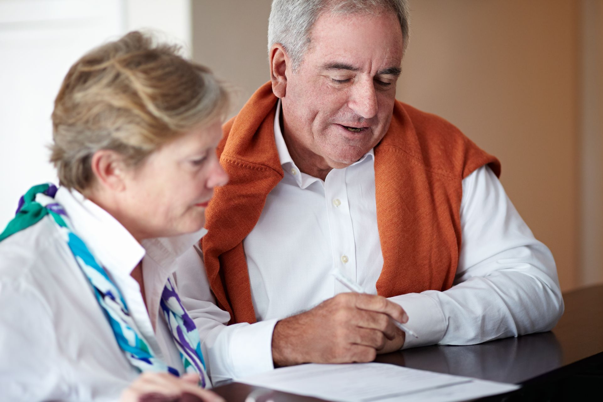 Two people in white shirts sit at a table, one writing on a document while the other looks on attentively.