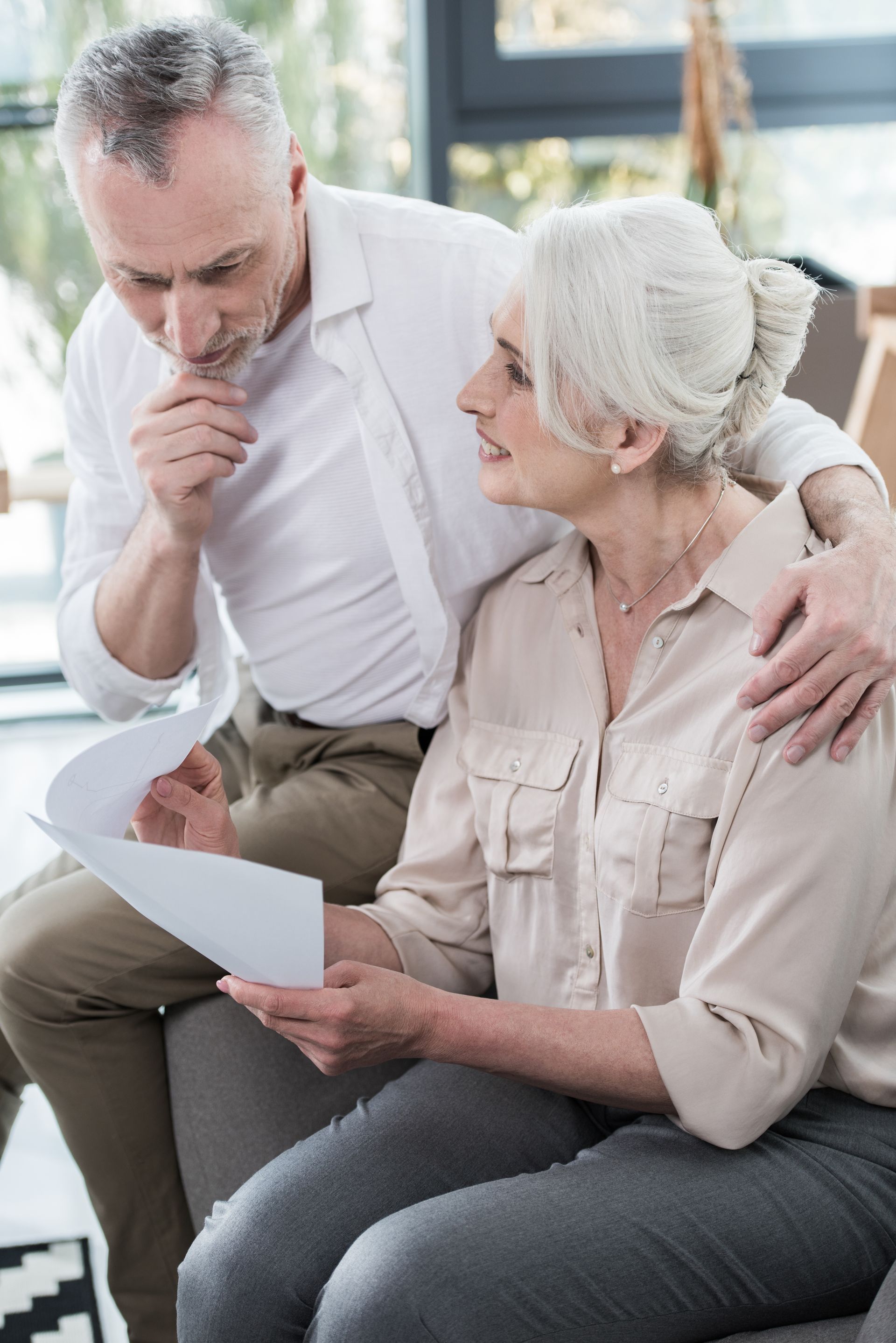 Two people sit on a couch reviewing documents, one looking thoughtful while the other looks on.