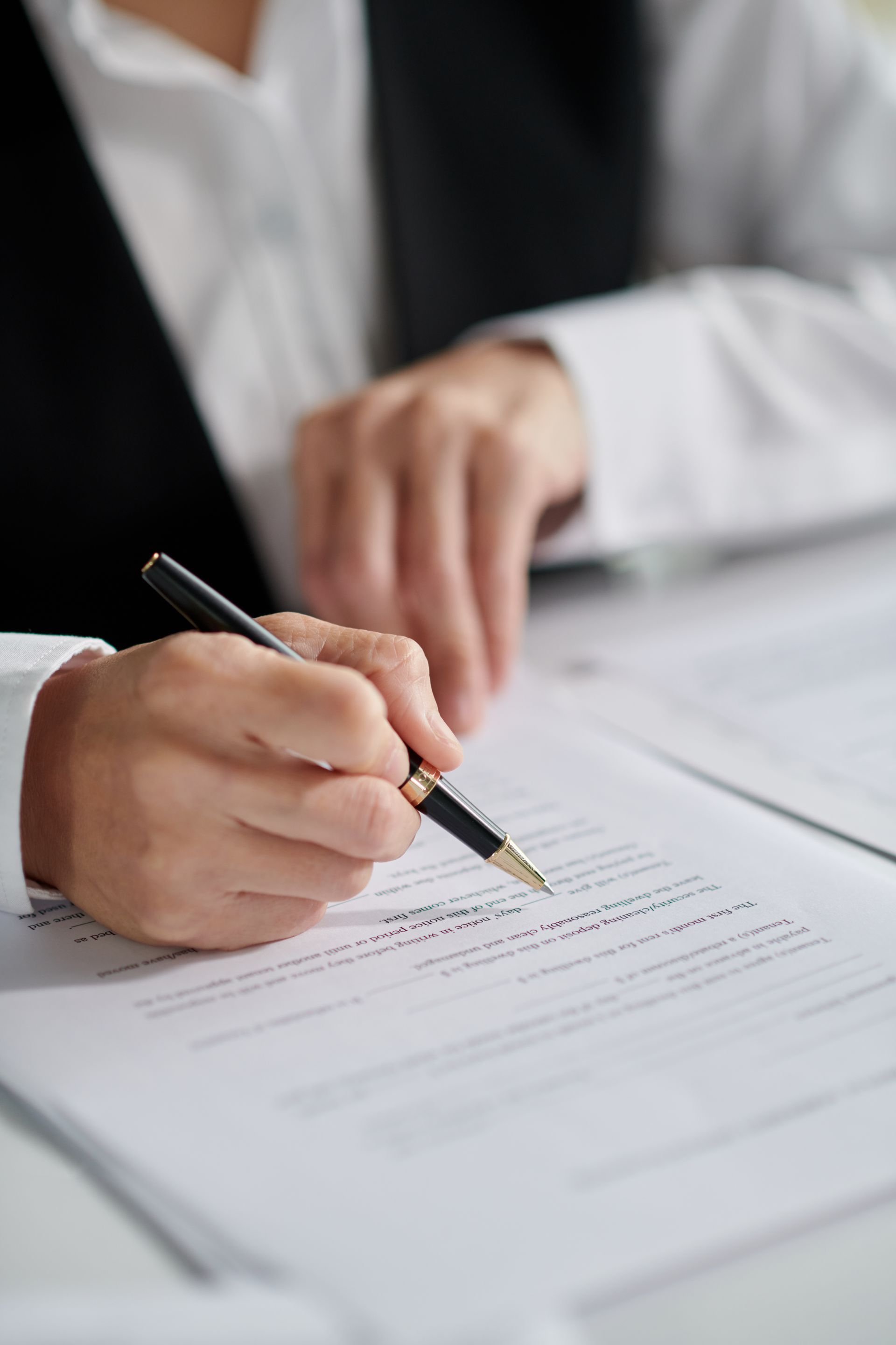 A close-up of a person's hand using a black pen to sign a formal document on a white desk.