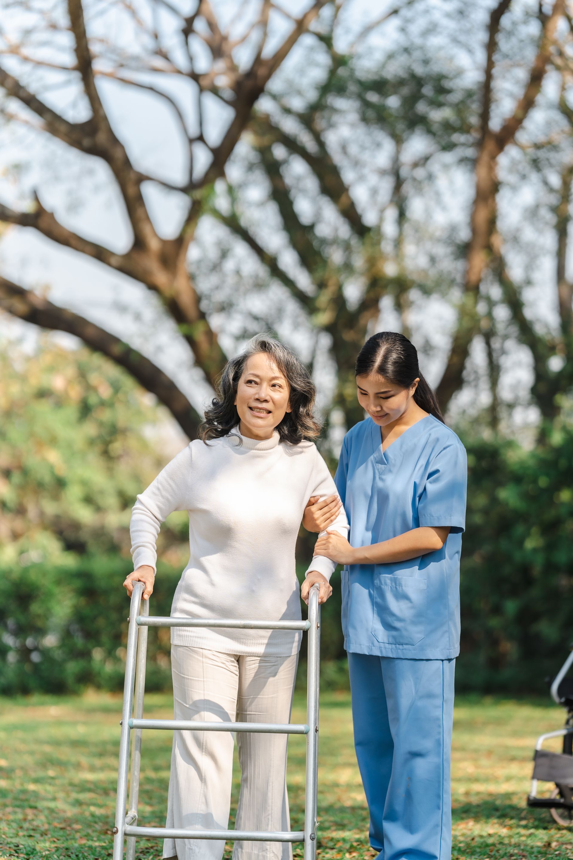 A caregiver in blue scrubs assists a person walking outdoors in a park using a silver metal walker.