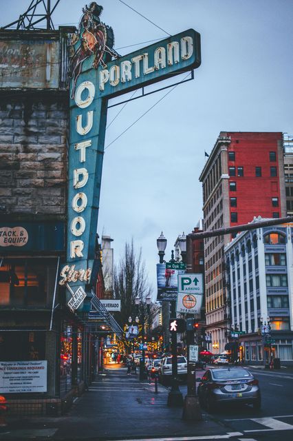 Downtown Portland street with the vertical “Portland Outdoor” sign at dusk, cars and city buildings in view