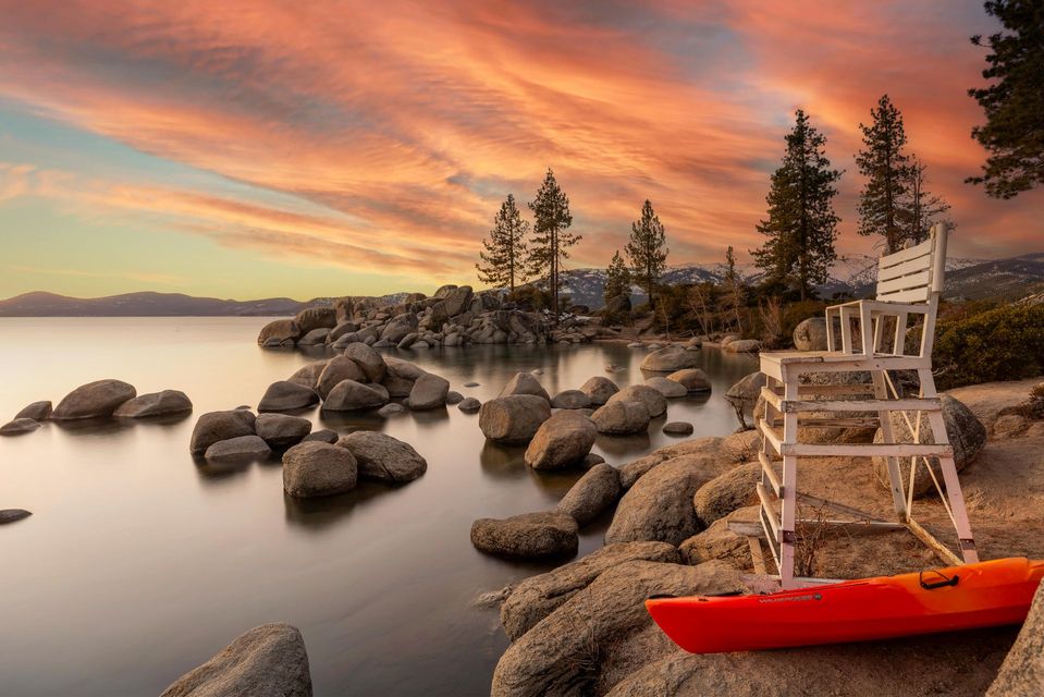 Rocky lakeshore at sunset with a red kayak, calm water, and a wooden chair on the shore