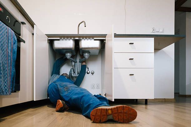 Plumber lying on the floor fixing complex dual-sink drainage pipes inside a kitchen cabinet.
