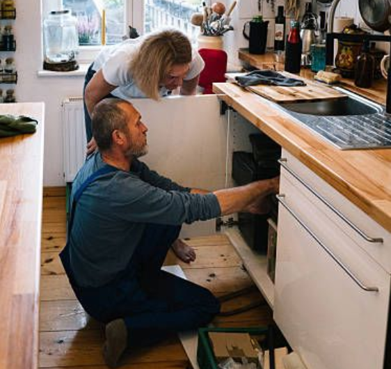 Residential plumber in overalls repairing an under-sink drainage system while a homeowner looks on.