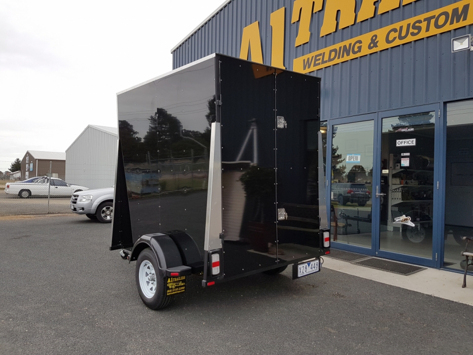 A black trailer is parked in front of a building that says welding and custom