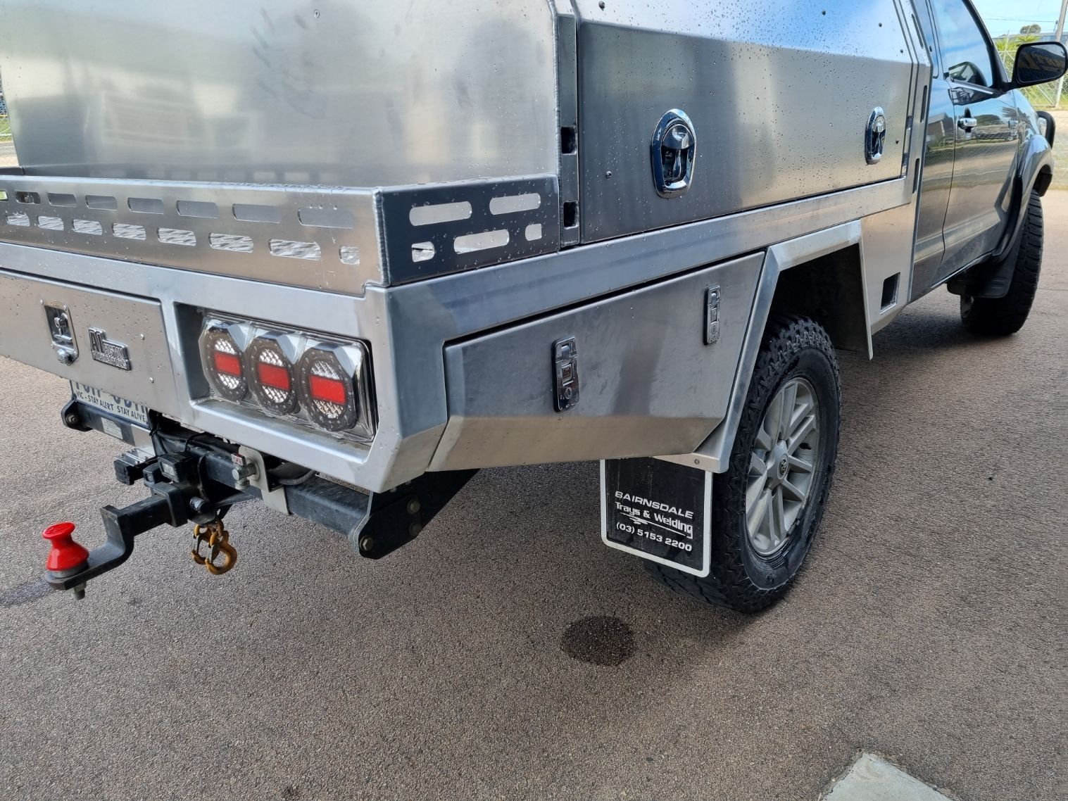 A silver utility truck with a custom metal tray, showing taillights, hitch, and mud flaps parked on pavement.