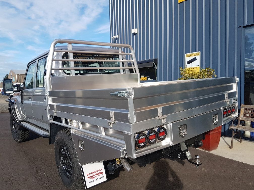 Silver pickup truck with an aluminum tray bed and a roll bar, parked outside a building with blue siding.