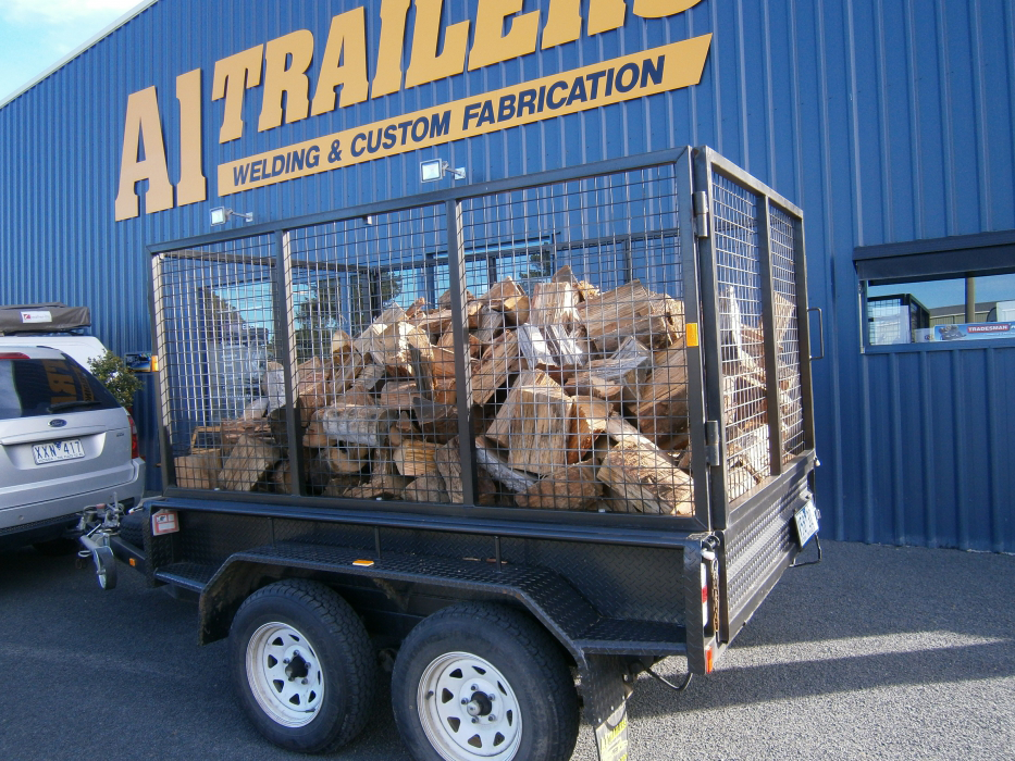 A trailer full of logs is parked in front of a welding and custom fabrication building