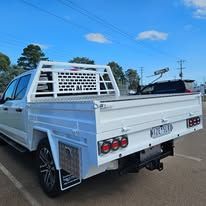 White pickup truck with a flatbed in an outdoor setting. The bed has a metal grille, and the truck is parked near a building under a blue sky.