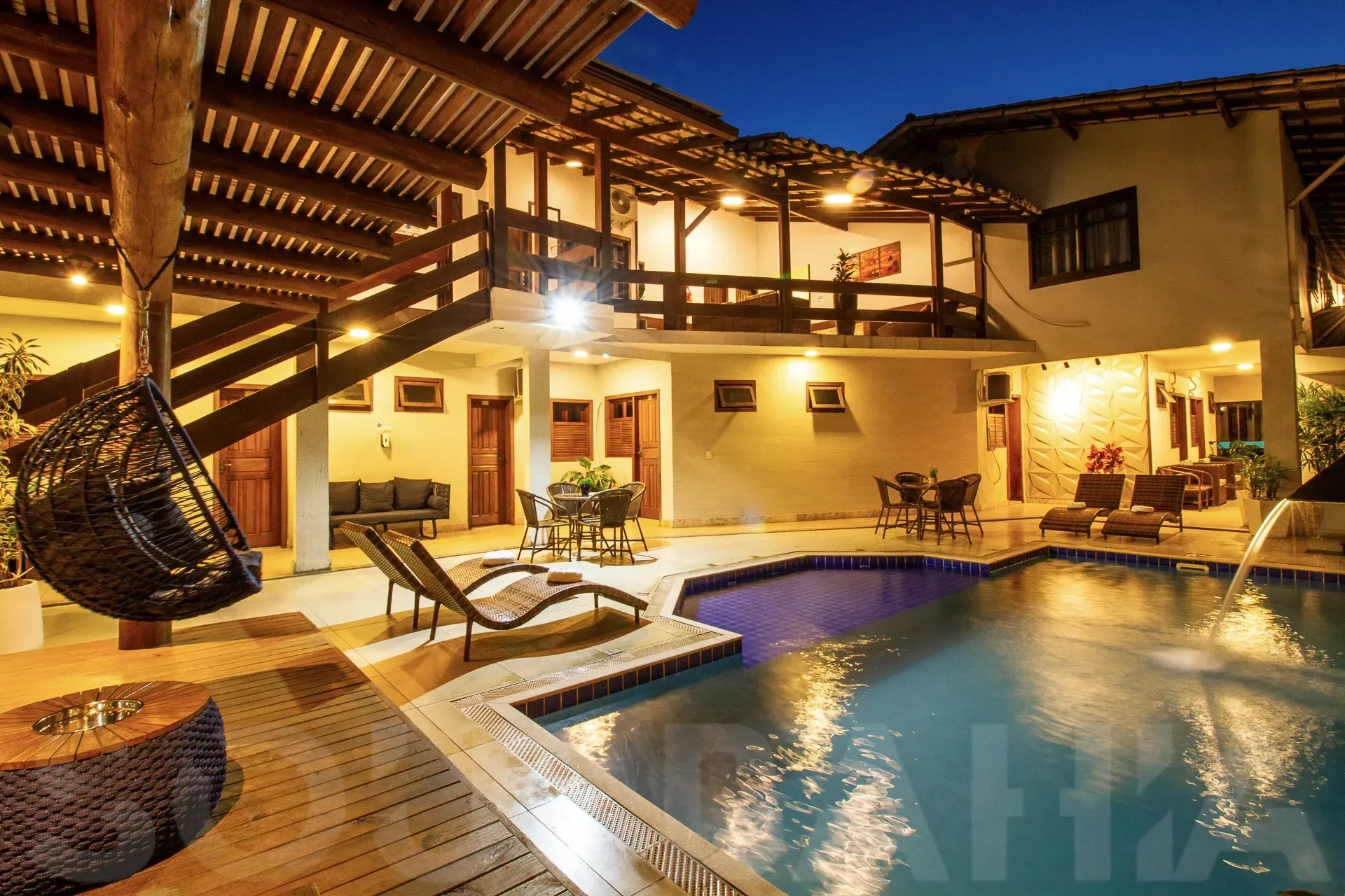 Pátio da piscina tropical iluminado à noite com espreguiçadeiras, deck de madeira e área de estar externa.
